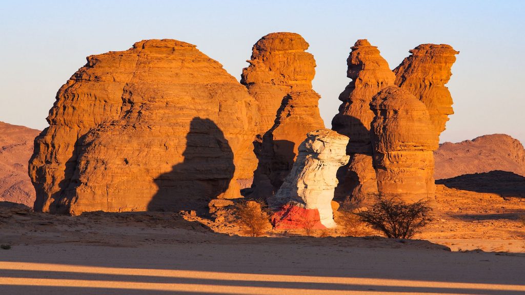 Tschad. Reisefotografie. Landschaftsfotografie. XL Digitaldruck auf Acryl Alu Dibond. Panoramadruck "Desert Art" zeigt Felsenformation, teilweise in rot und weiß bemalt. Tamarisken im Vordergrund. Licht-und Schatten. Reisefotografie. Chad Republic. XL digital print on acrylic alu dibond. Panoramic image "Desert Art" shows rock formations partially painted in red and white. in the foreground tamarisk trees. light and shadow. travel photography. landscape photography.